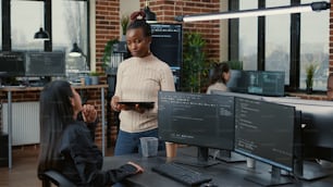 Software developer standing up holding digital tablet talking with programer sitting at desk with computer screens showing running code. System engineer coding with team writing machine learning data.