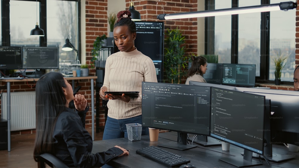 소프트웨어 개발자는 디지털 태블릿을 들고 서서 컴퓨터 화면에 실행 중인 코드가 표시된 책상에 앉아 있는 프로그래머와 이야기합니다. 기계 학습 데이터를 작성하는 팀과 함께 코딩하는 시스템 엔지니어.