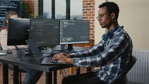 Programer thinking while touching beard and fixing glasses while typing on laptop sitting at desk with multiple screens parsing code. Focused database admin working with team coding in the background.