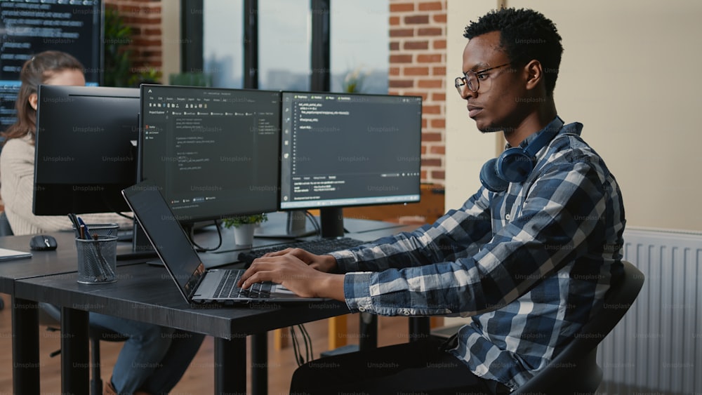 Programer thinking while touching beard and fixing glasses while typing on laptop sitting at desk with multiple screens parsing code. Focused database admin working with team coding in the background.