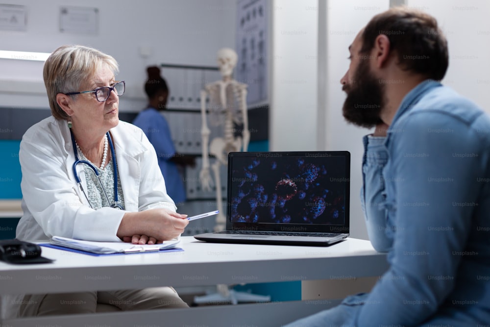 Medico che spiega l'illustrazione dei batteri del coronavirus sul laptop all'uomo nel gabinetto. Medico e paziente che guardano lo schermo con animazione del virus per comprendere la malattia covid 19 e la diagnosi.
