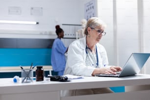 Médico sênior usando laptop no armário para procurar tratamento de saúde. Clínico geral olhando para a tela do computador para trabalhar em medicamentos prescritos contra o diagnóstico de doenças.