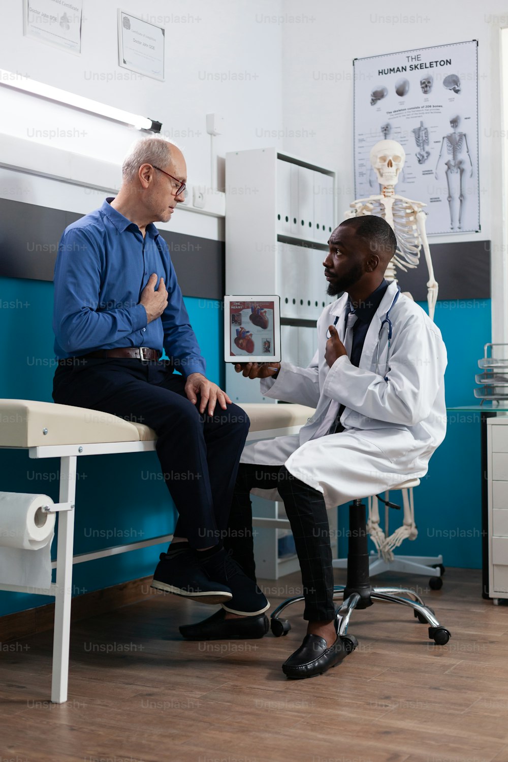 Afroamerikanischer Therapeut, Arzt, der einen Tablet-Computer mit Herz-Radiographie hält und dem pensionierten alten Patienten während eines klinischen Termins im Krankenhausbüro das Fachwissen über Krankheiten erklärt. Medizin-Konzept