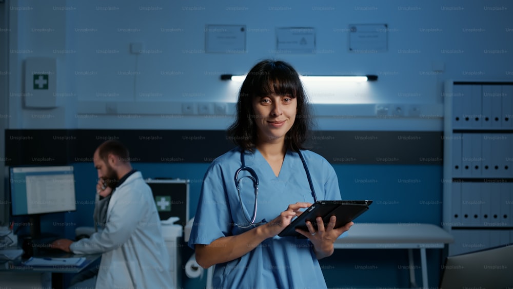 Portrait of medical assistant checking disease expertise on laptop computer working in hospital office after hours. Physician nurse with stethoscope typing healthcare treatment. Medicine concept