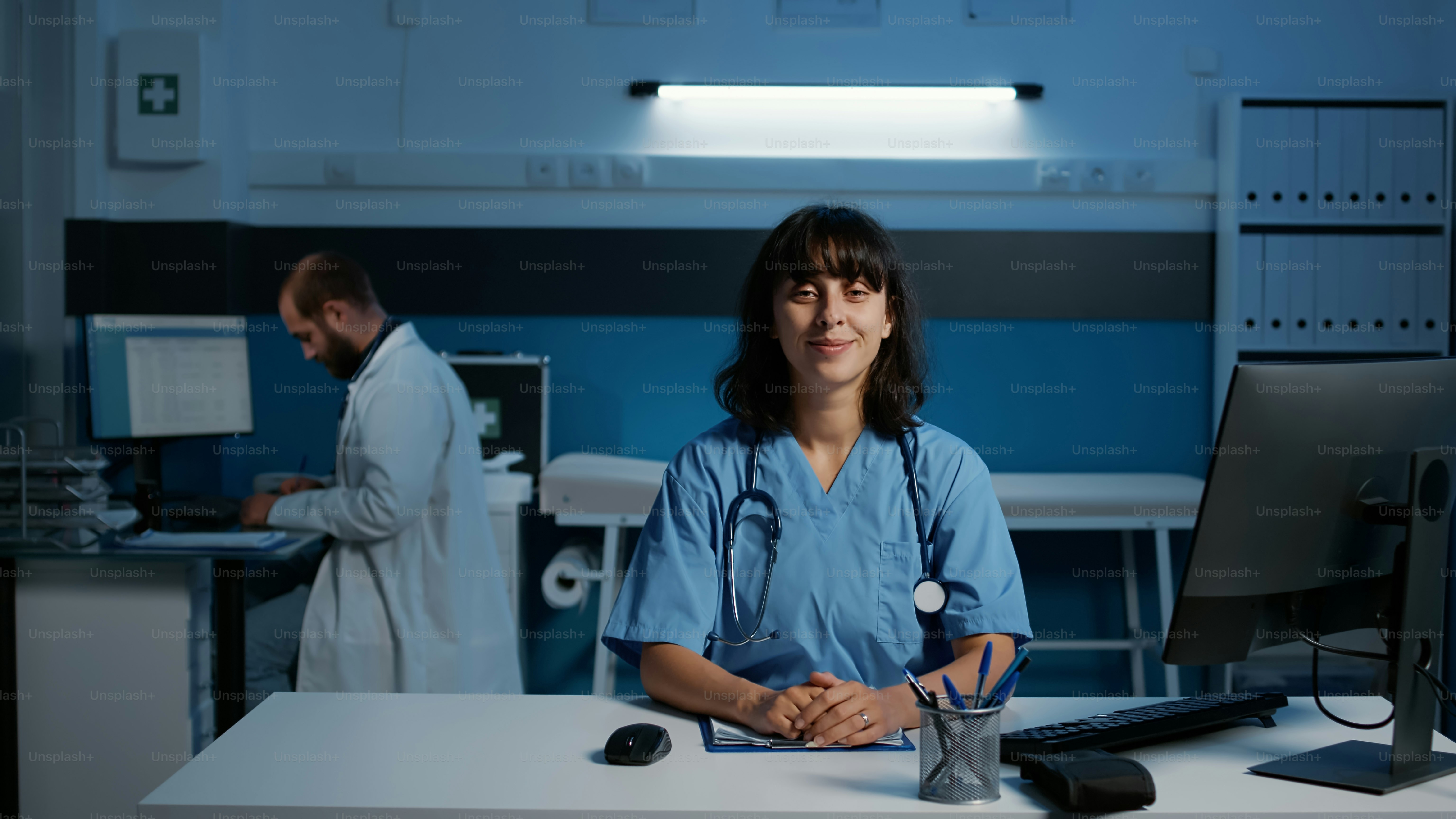 Lächelnde Arzthelferin, die am Schreibtischtisch steht und Krankheitssymptome auf dem Computer überprüft, Gesundheitsbehandlungen eintippt. Medizinische Fachangestellte, die während der Konsultation nach Feierabend im Krankenhausbüro arbeitet