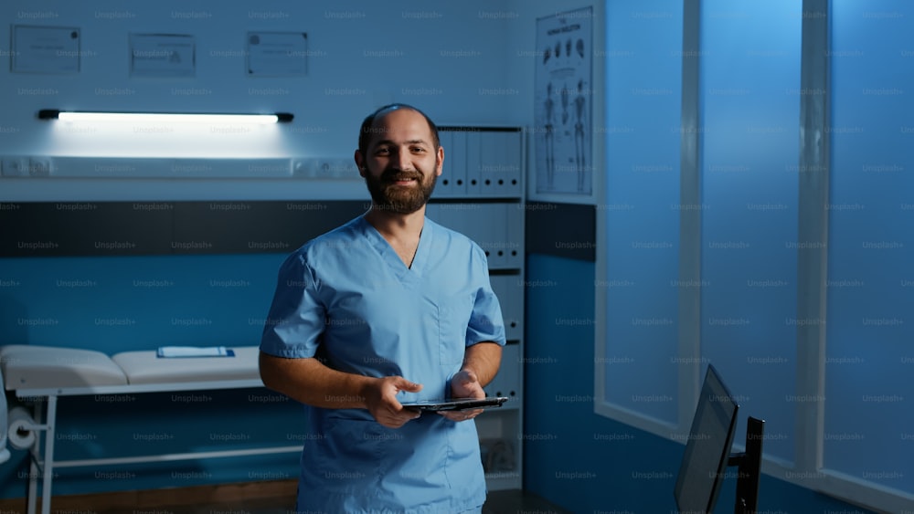Caucasian man assistant holding tablet computer typing medical expertise checking patient disease report. Nurse in blue uniform working after hours in hospital office. Health care service and concept