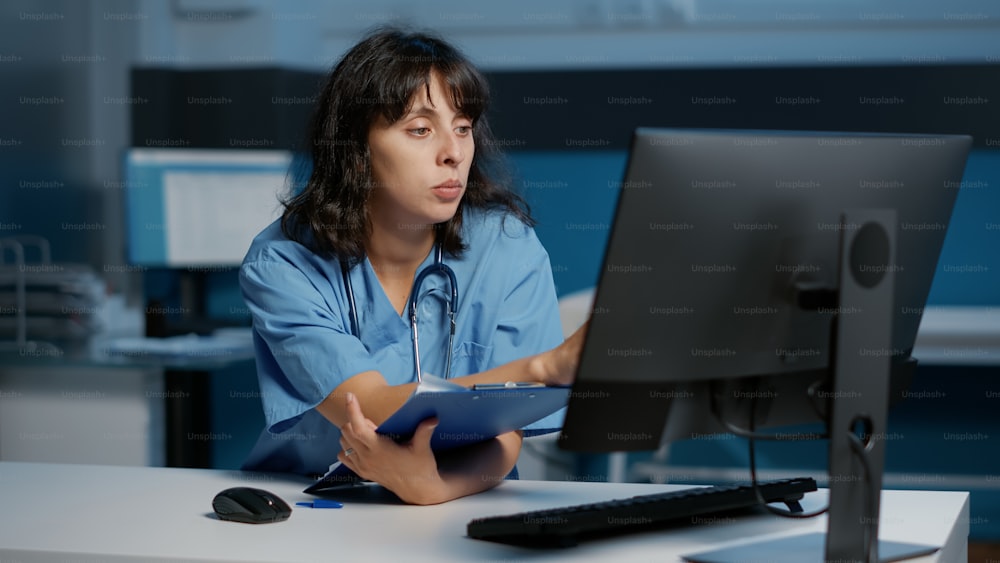 Die Krankenschwester überprüft den Krankheitsbericht auf dem Computer, während sie an der Patientenbehandlung arbeitet, um die Krankheit während der Nachtschicht im Krankenhausbüro zu heilen. Medizinische Fachangestellte, die Papiere mit Fachwissen betrachtet