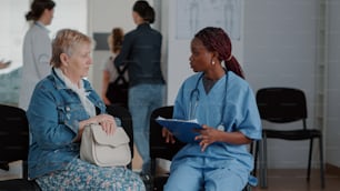 Enfermeira afro-americana explicando o diagnóstico para idosa em sala de espera no ambulatório. Assistente médico e paciente com doença falando sobre tratamento de saúde e recuperação.