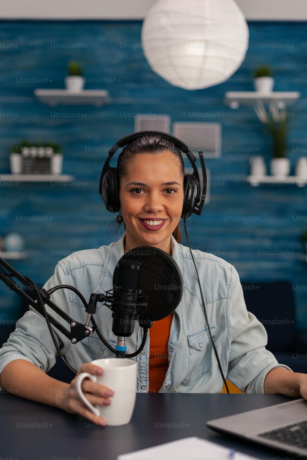 Portait of woman vlogger holding cup and streaming podcast using laptop. Social media influencer recording livestream with headphones and microphone in home office. Smiling blogger podcasting.