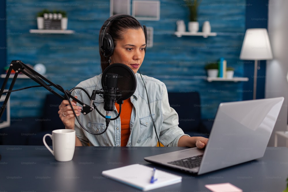 Woman vlogger streaming podcast and using laptop at desk. Influencer with headphones in home office recording livestream on computer. Blogger live broadcasting for social media with microphone.