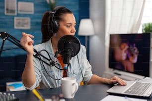 Woman talking on microphone while using laptop. Content creator wearing headphones recording podcast with audio equipment. Social media influencer in home office broadcasting live.