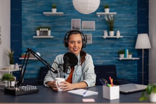 Portrait of social media influencer wearig headphones and streaming podcast. Content creator recording broadcast with headphones and microphone in home office. Smiling woman vlogger podcasting.