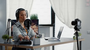 Creative content creator recording vlog in home studio while tweaking knobs on audio mixer. Internet live broadcast talk show host talking with audience while using professional audio equipment.