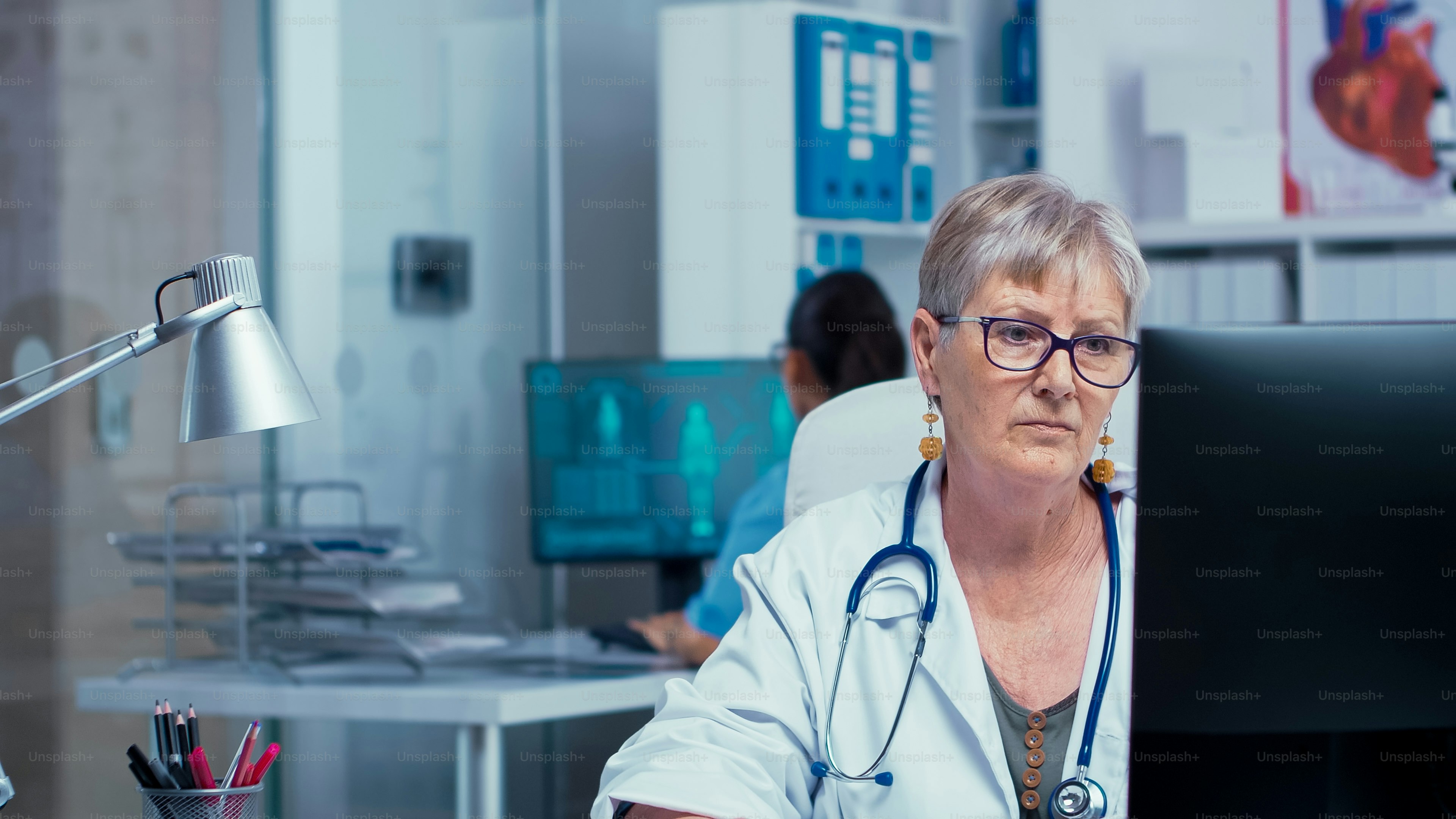 Portrait of senior doctor woman working on PC in modern private cabinet while the nurse is checking X Rays in the back and other medical stuff walks in hallway. Consulting in healthcare system