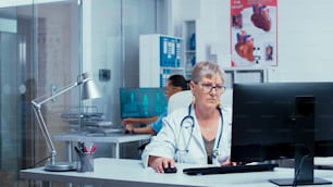 Authentische erfahrene Oberärztin, die am PC in einer modernen Privatklinik mit Glaswänden arbeitet, Patientin im Rollstuhl im Hintergrund und anderes medizinisches Personal, das durch den Flur geht. Medizin und Gesundheitssystem