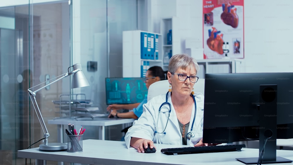 Authentic experienced senior woman doctor working on PC in modern private clinic with glass walls, patient in wheelchair in background and other medical staff going through hallway. Medicine and healthcare system
