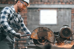 Cutting the metal by saw. Factory male worker in uniform is indoors.