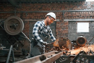 제복을 입고 안전모를 쓴 공장 남성 노동자가 실내에서 금속을 자르고 있다.