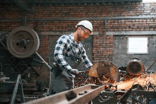 Factory male worker in uniform and hard hat is indoors, cutting the metal.