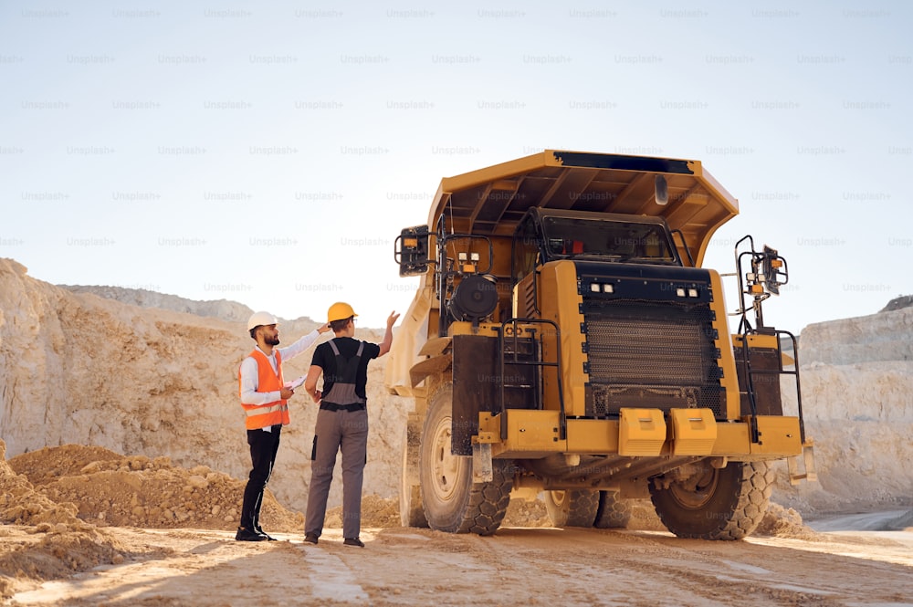 Standing near haul truck and talking about document. Two men in uniform are working together in the quarry.