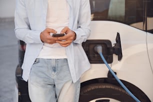 De pé com o smartphone enquanto o automóvel está carregando. Vista de perto do homem com seu carro elétrico.