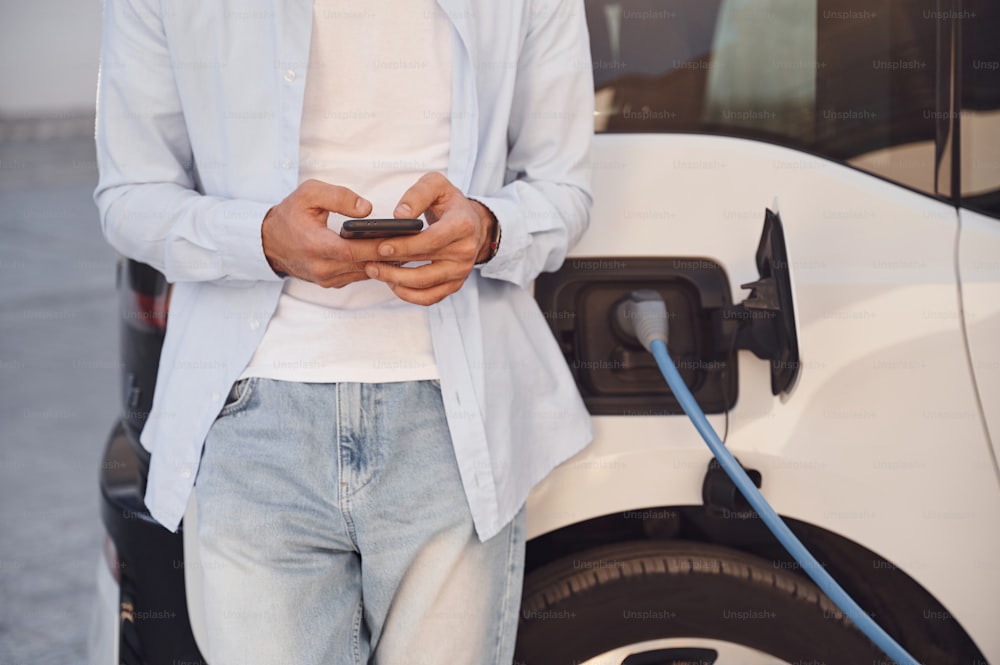 Debout avec un smartphone pendant que l’automobile est en charge. Vue rapprochée de l’homme avec sa voiture électrique.