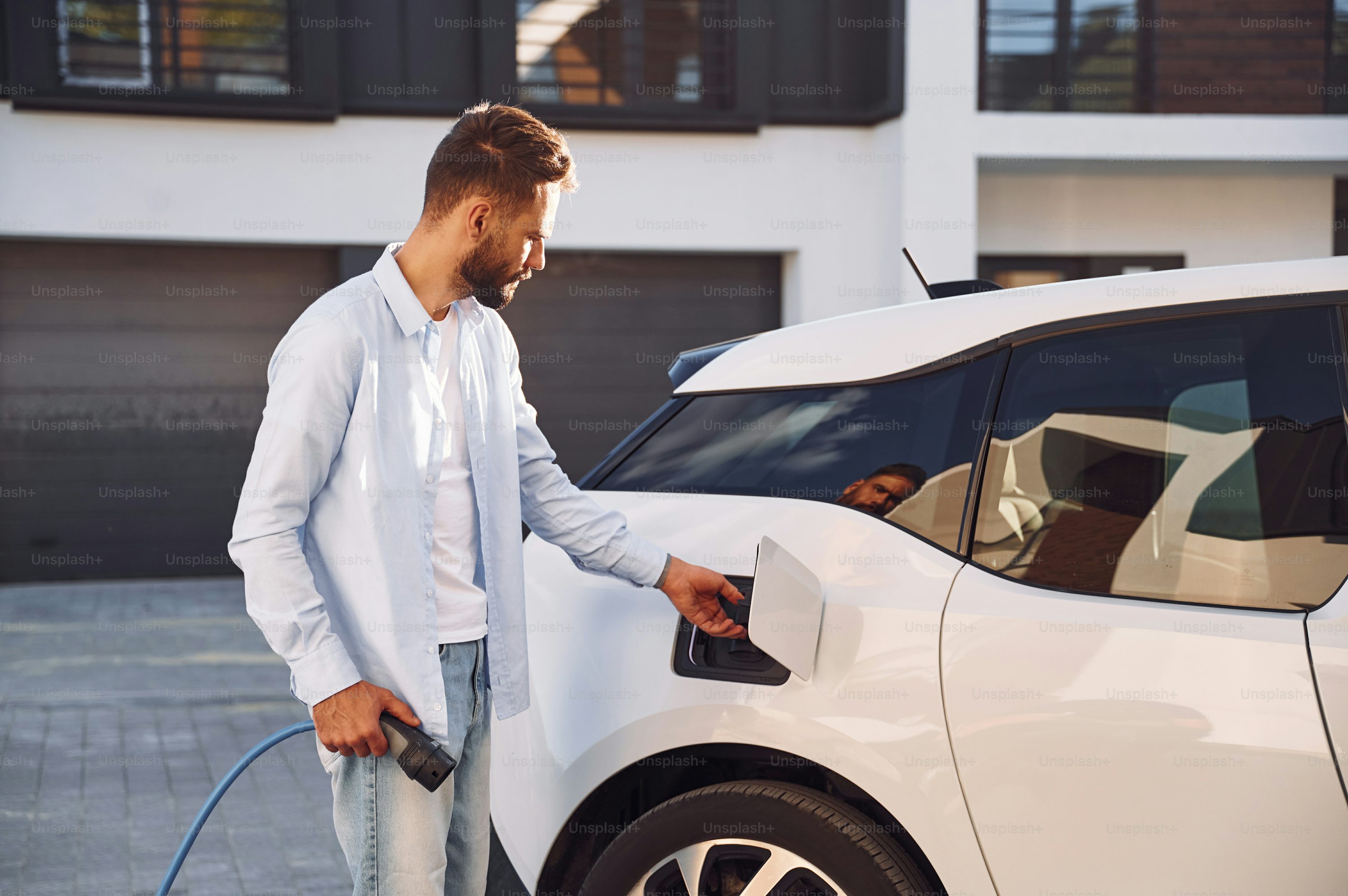 Plugging in charging cable. Young stylish man is with electric car at daytime.