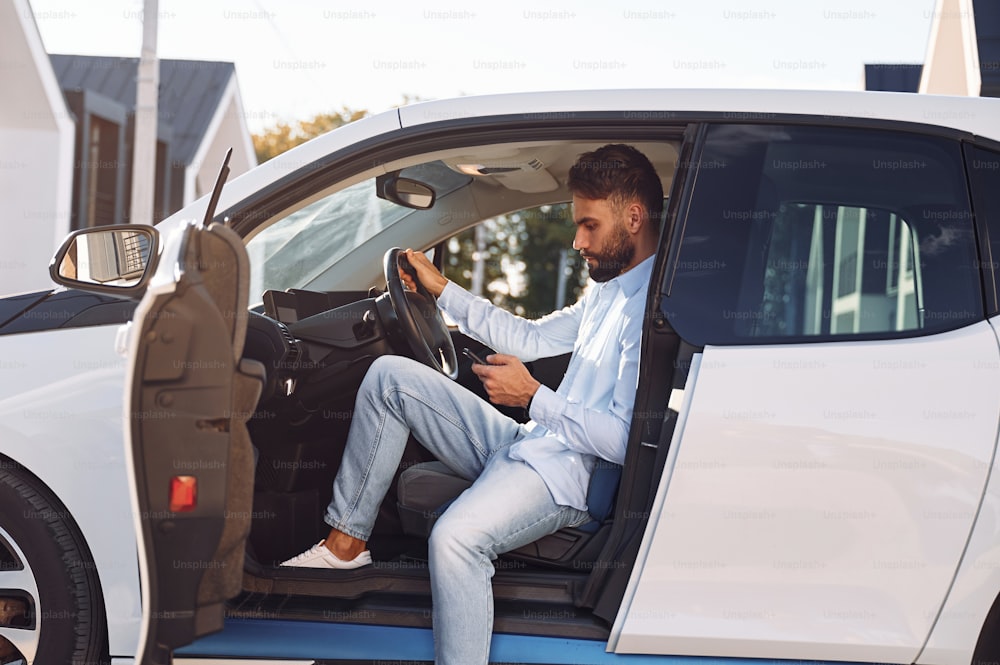 Assis dans le véhicule. Un jeune homme élégant est avec une voiture électrique pendant la journée.