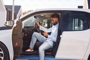 Sentado en el vehículo. Joven con estilo está con coche eléctrico durante el día.