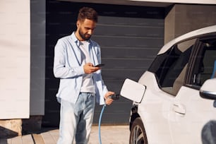 Sunny weather. Holding smartphone and charging the vehicle. Young stylish man is with electric car at daytime.