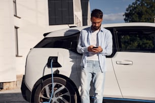 Tempo ensolarado. Segurando o smartphone e carregando o veículo. O jovem homem elegante está com carro elétrico durante o dia.