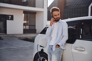 Parler au téléphone. Un jeune homme élégant est avec une voiture électrique pendant la journée.