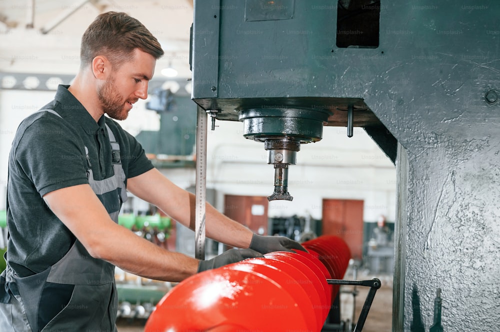 Measuring red colored sharp part. Man in uniform is in workstation developing details of agriculture technique.