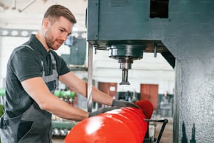 Measuring red colored sharp part. Man in uniform is in workstation developing details of agriculture technique.