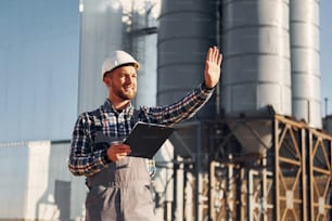 In grey clothes. Construction worker in uniform is outdoors near the factory.