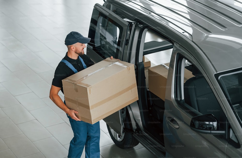 Walking and holding box in hands. Delivery man in uniform is indoors with car and with order.