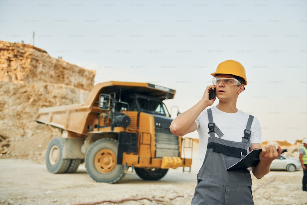 In glasses and hard hat. Worker in professional uniform is on the borrow pit at daytime.