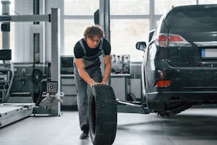 Works with tire. Adult man in grey colored uniform is in the automobile salon.