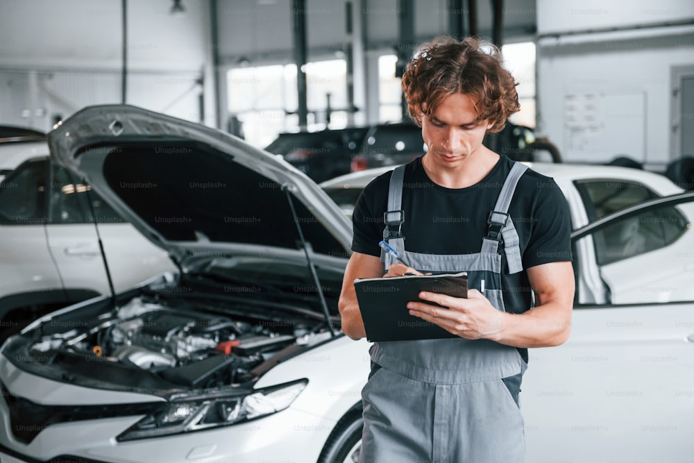 With notepad in hands. Adult man in grey colored uniform works in the automobile salon.