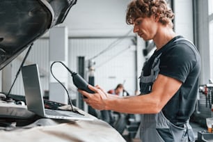 Special tool for testing characteristics. Adult man in grey colored uniform works in the automobile salon.