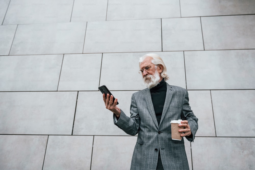 Utilisation d’un smartphone. L’homme d’affaires senior en vêtements formels, avec les cheveux gris et la barbe est à l’extérieur.