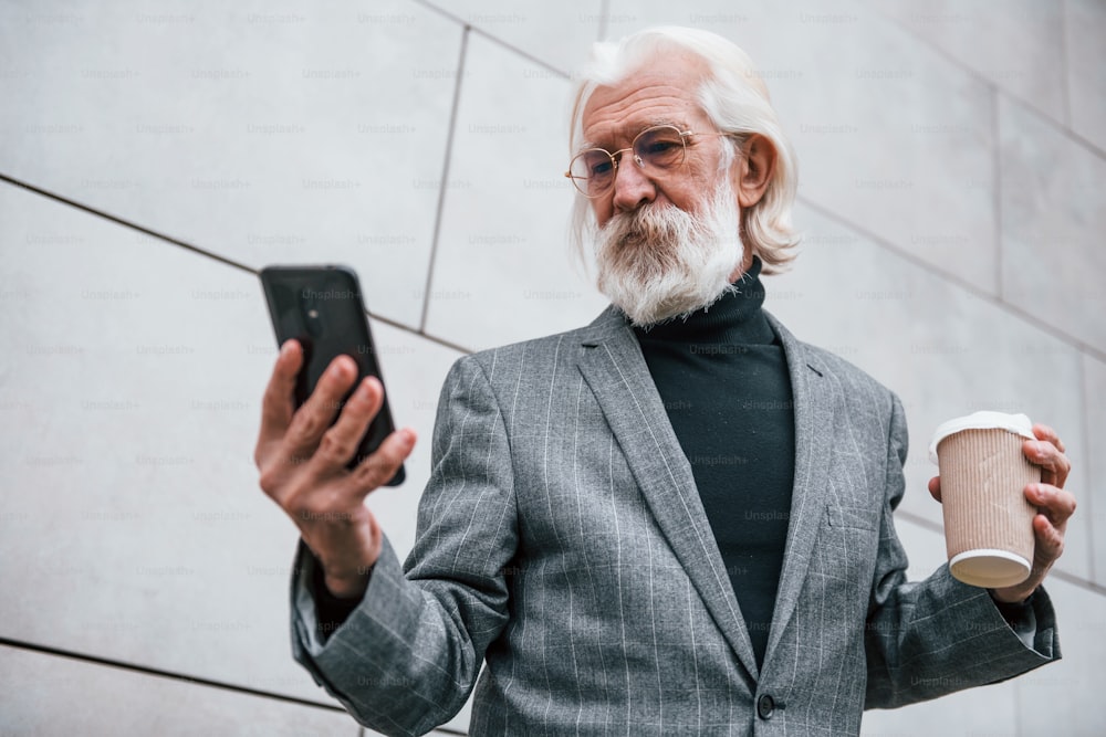 Mit dem Smartphone. Älterer Geschäftsmann in formeller Kleidung, mit grauen Haaren und Bart ist im Freien.