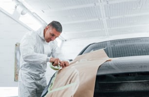 Covering car by brown cloth. Caucasian automobile repairman in uniform works in garage.