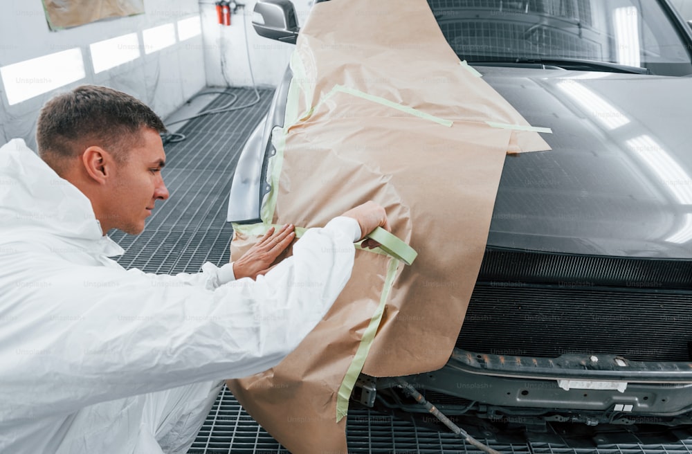 Covering car by brown cloth. Caucasian automobile repairman in uniform works in garage.