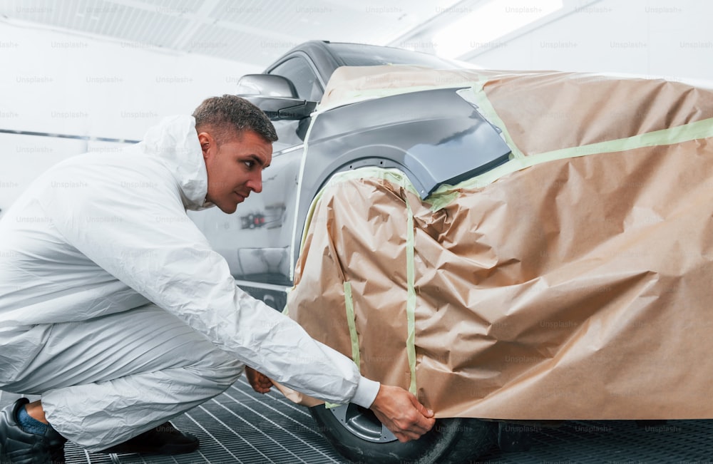 Covering car by brown cloth. Caucasian automobile repairman in uniform works in garage.