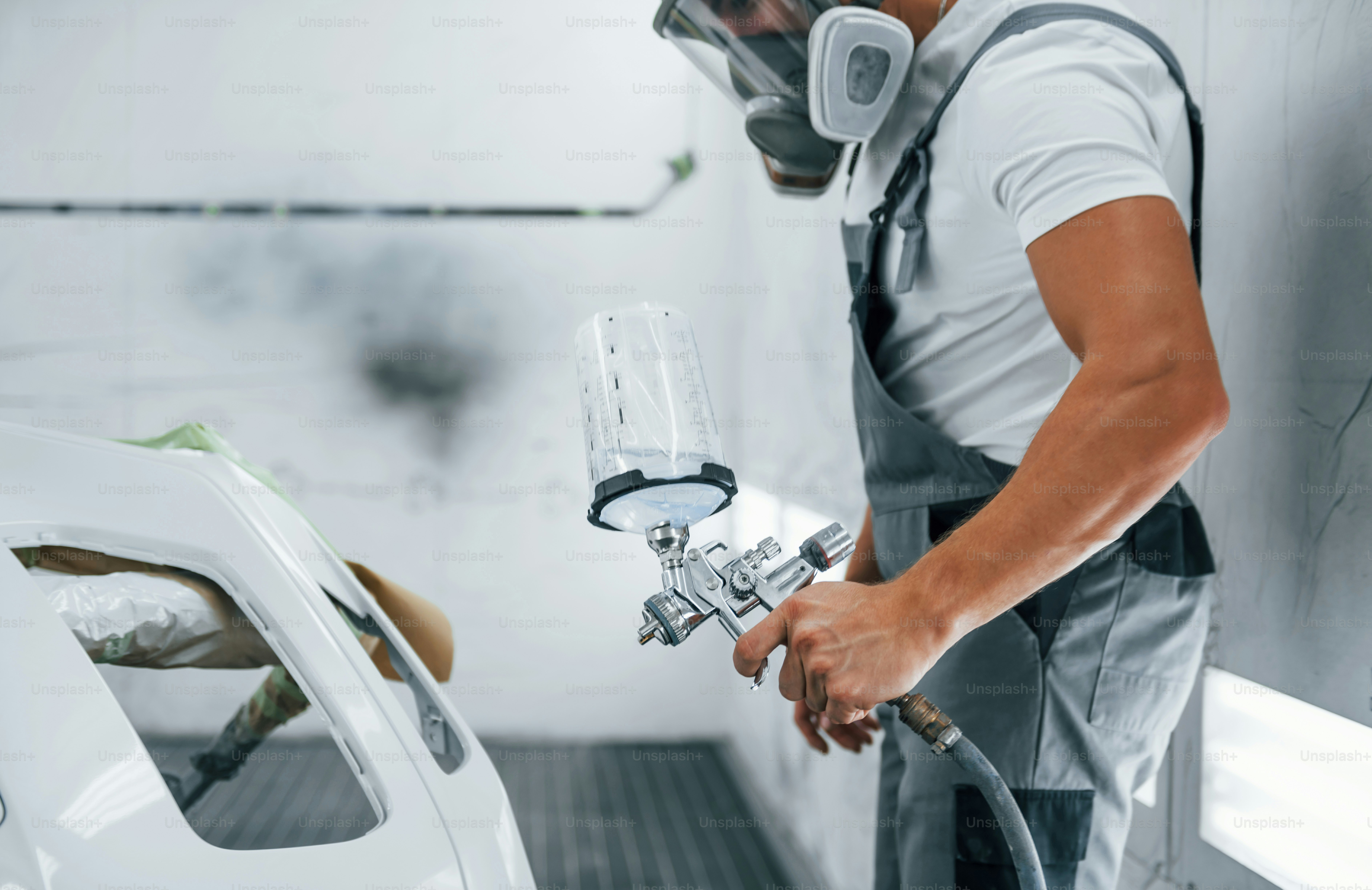 With painting gun. Caucasian automobile repairman in uniform works in garage.