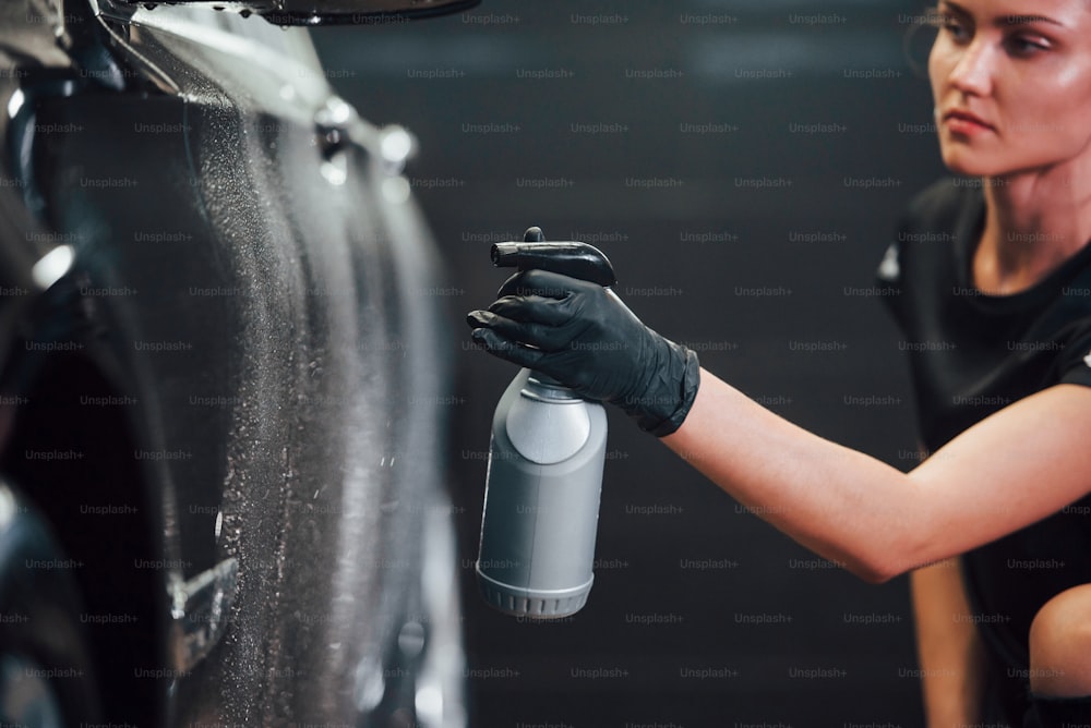 Spraying the vehicle. Modern black automobile get cleaned by woman inside of car wash station.
