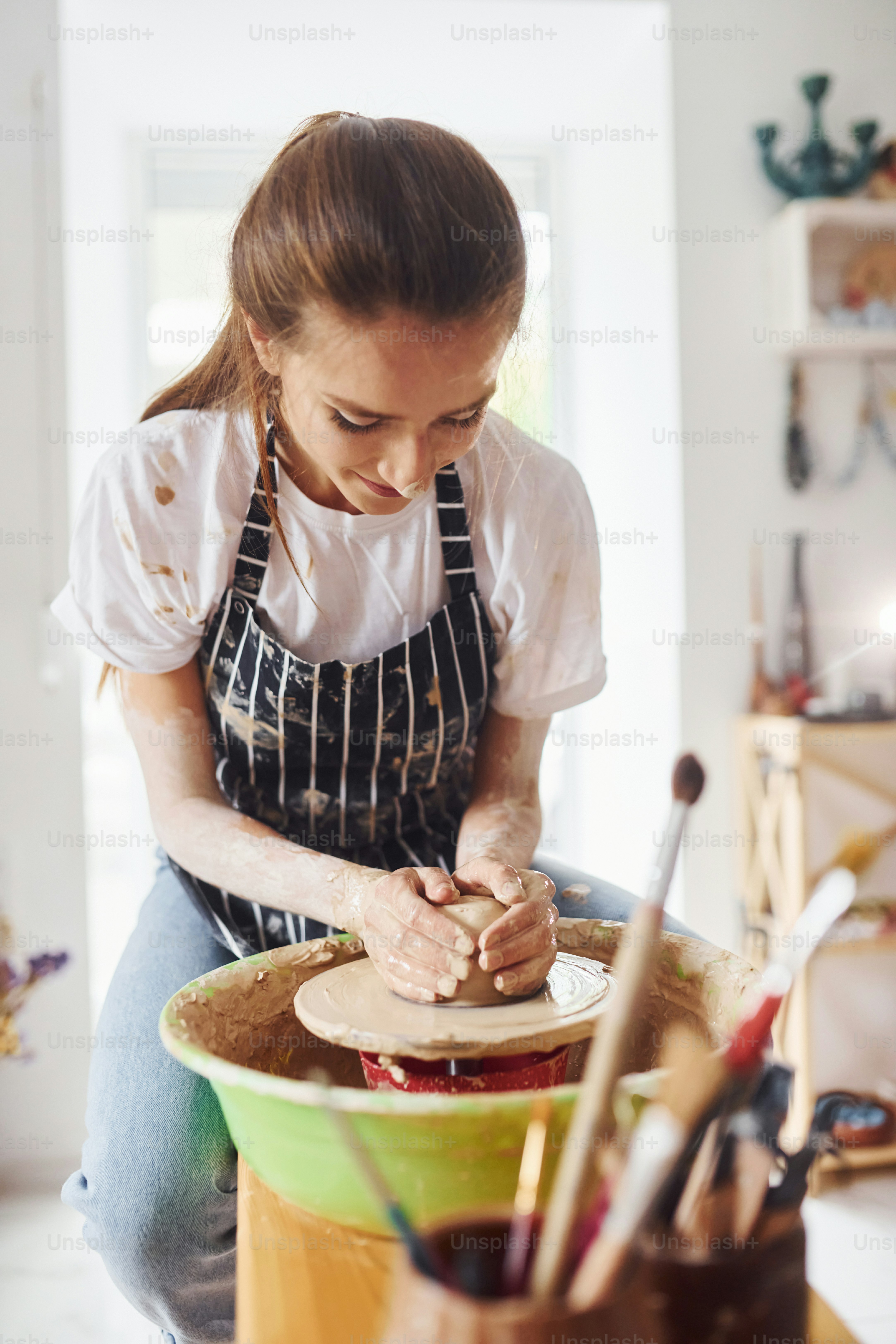 Young female ceramist working by using pottery wheel indoors and making handmade clay product.