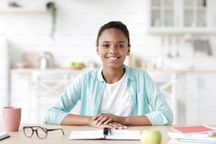 Modern pupil study at home, lesson and class remotely, new normal and be safe. Headshot of smiling teen african american girl looking at camera at table with notebooks in kitchen interior, free space