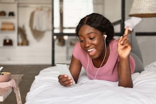 Cheerful African American Female Listening To Music On Phone Wearing Earbuds Earphones Lying On Bed At Home. Millennial Lady Singing Song Holding Smartphone Relaxing In Bedroom
